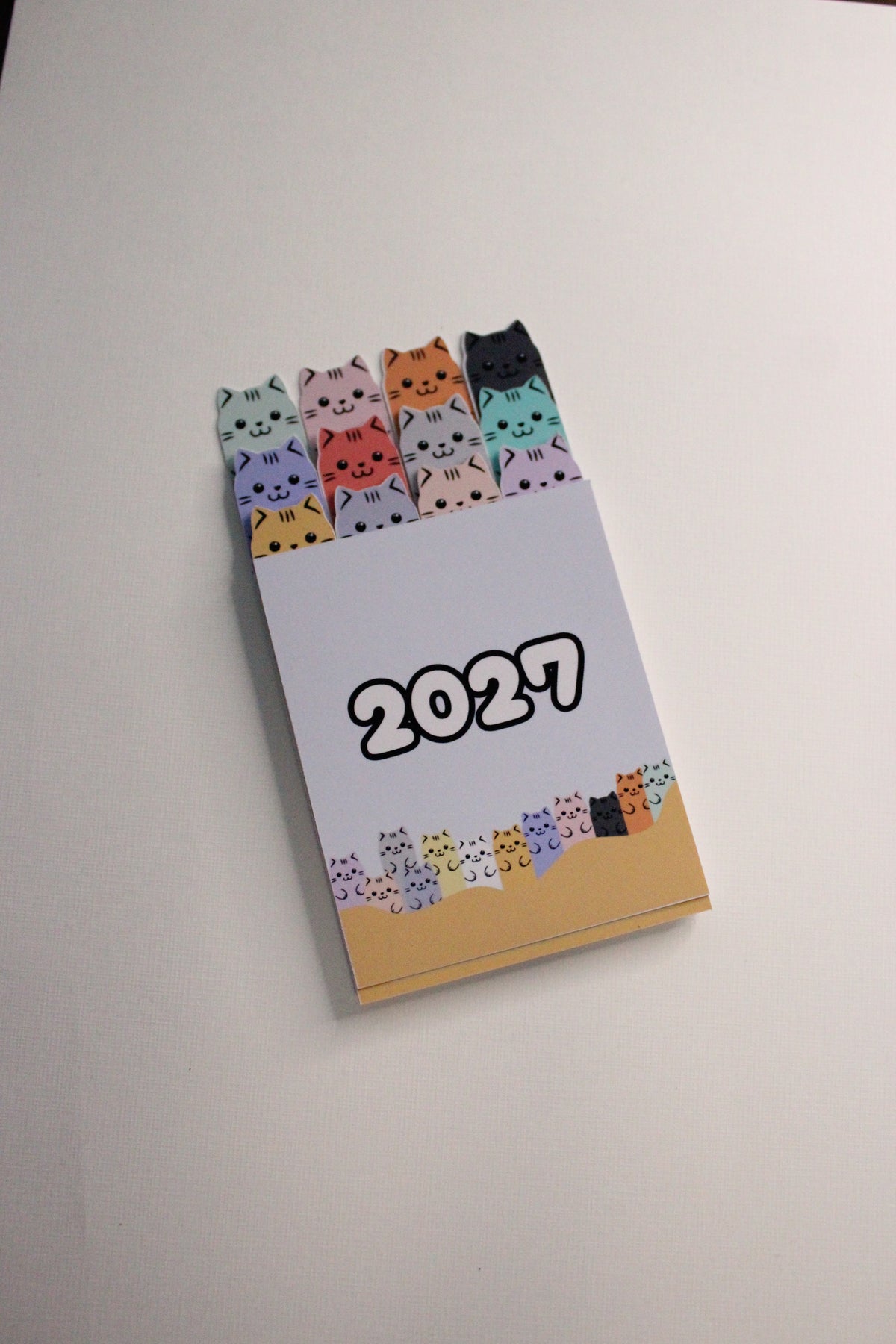 A 2027 pocket calendar with cute cartoon cat illustrations, shown partially open on a light gray surface.